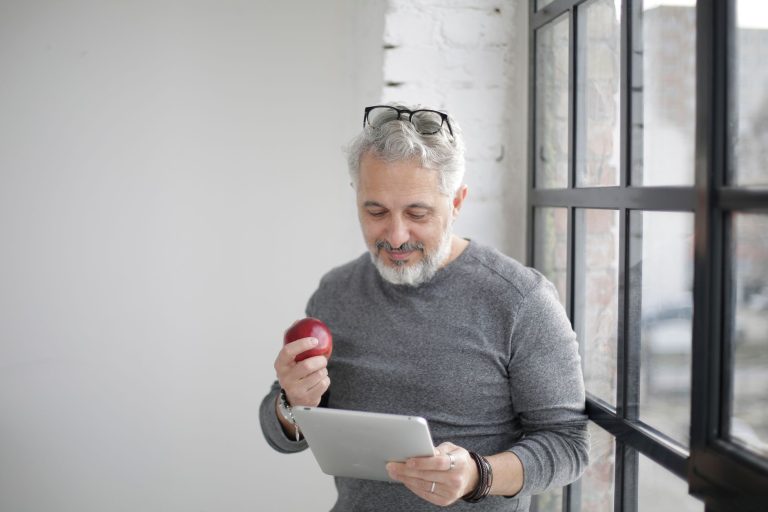 Middle aged bearded man in casual wear with eyeglasses on head watching video on tablet while having break with apple near window