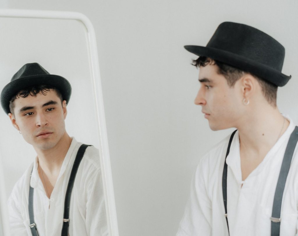 A stylish man in a hat and suspenders looks at himself in the mirror.