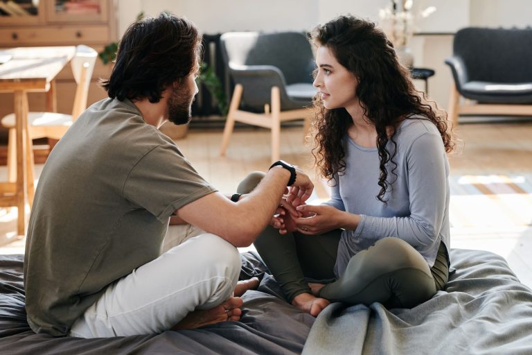 A couple sitting closely on a bed, engaged in a thoughtful discussion in a cozy home setting.