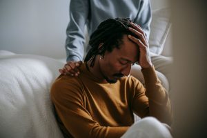 0e7e75c2 55e1 4f35 8afd 084575919a1d Worried depressed African American male touching head nervously and sitting with eyes closed near supporting wife in bedroom