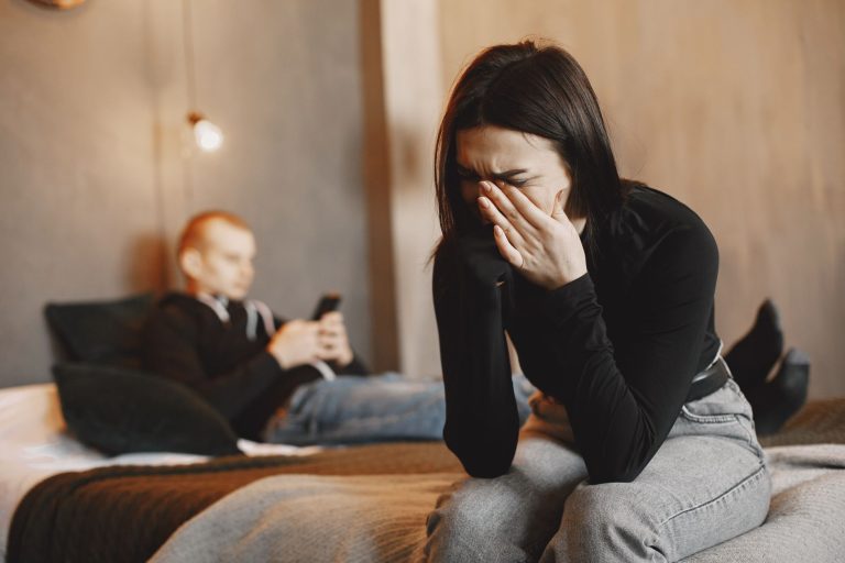 8c5460a0 c0c7 463f 99ac 1b421cd28d14 Woman crying on bed, man in background, illustrating relationship stress.