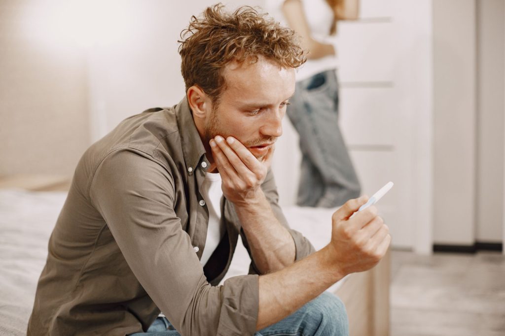 A concerned man sitting on a bed, holding a pregnancy test, deep in thought.