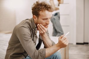 A concerned man sitting on a bed, holding a pregnancy test, deep in thought.