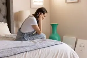 Side view of a woman sitting thoughtfully on a bed in a cozy room, lost in contemplation.