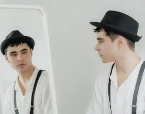 A stylish man in a hat and suspenders looks at himself in the mirror.