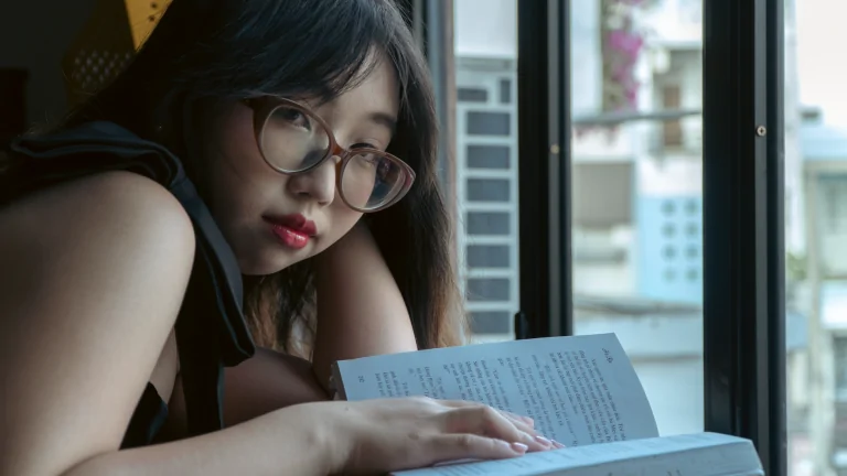A young woman with glasses reads a book by a window on a calm day.